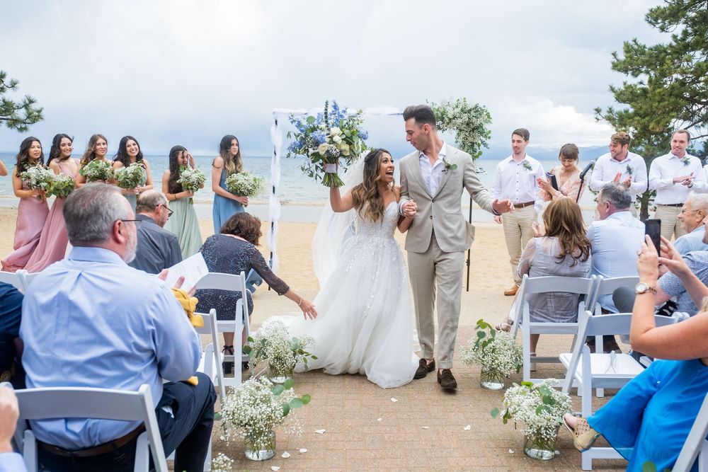 A joyful wedding ceremony takes place outdoors on a hilltop with guests seated in white chairs and blue accents.