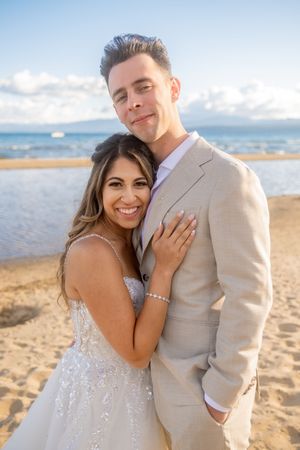 Newlywed couple embracing on a beach at sunset with mountains and clouds in the background.