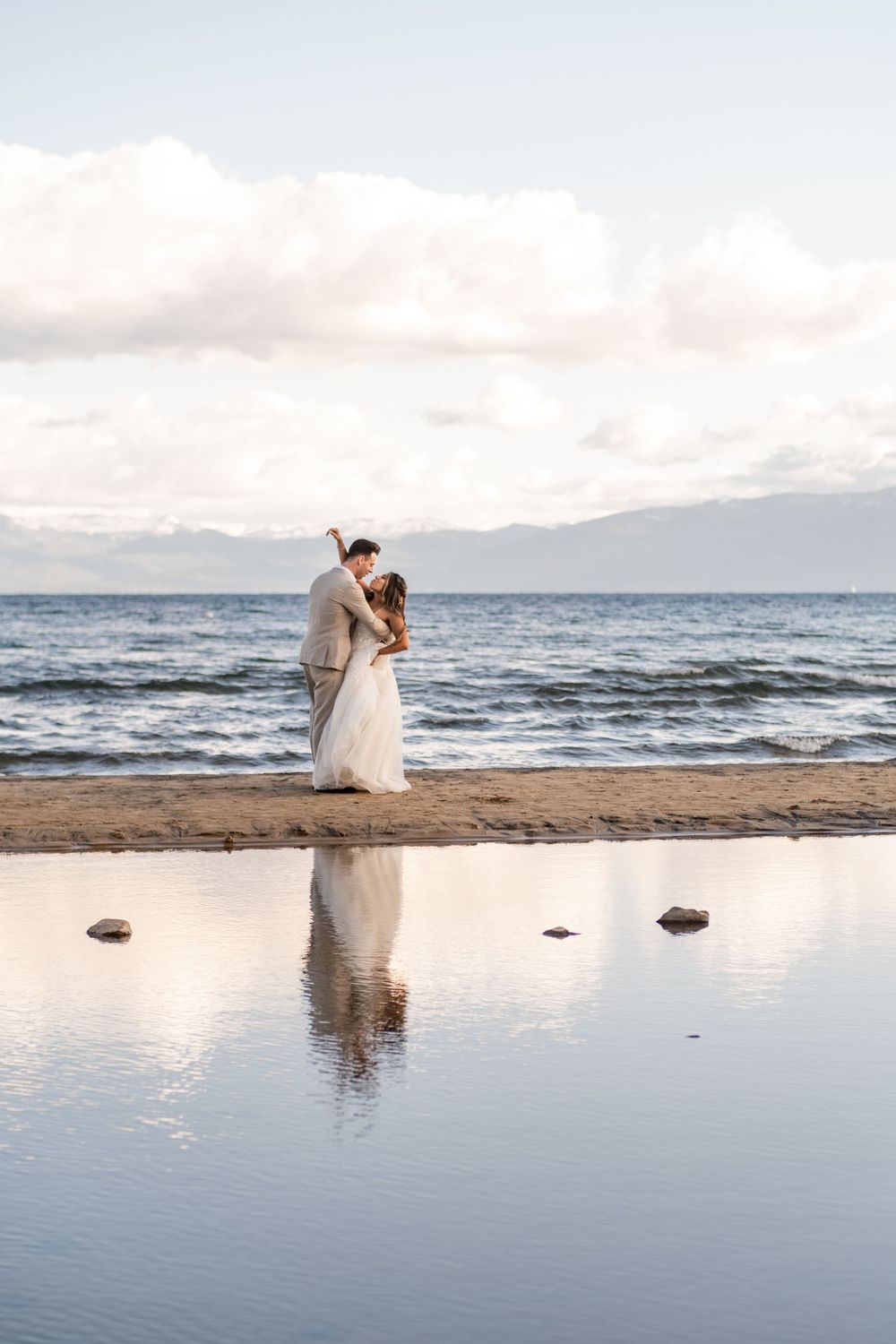 A romantic couple shares a tender moment along a serene beach shoreline at sunset with waves gently rolling in.