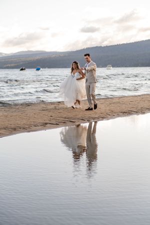 A couple walks along a beach at sunset with mountains in background and their reflection mirrored in calm water.