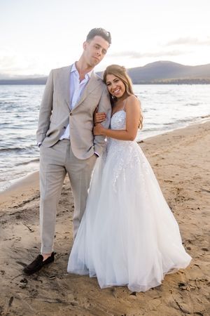 Newlywed couple in a gray suit and white wedding dress embrace on a scenic beach at sunset.