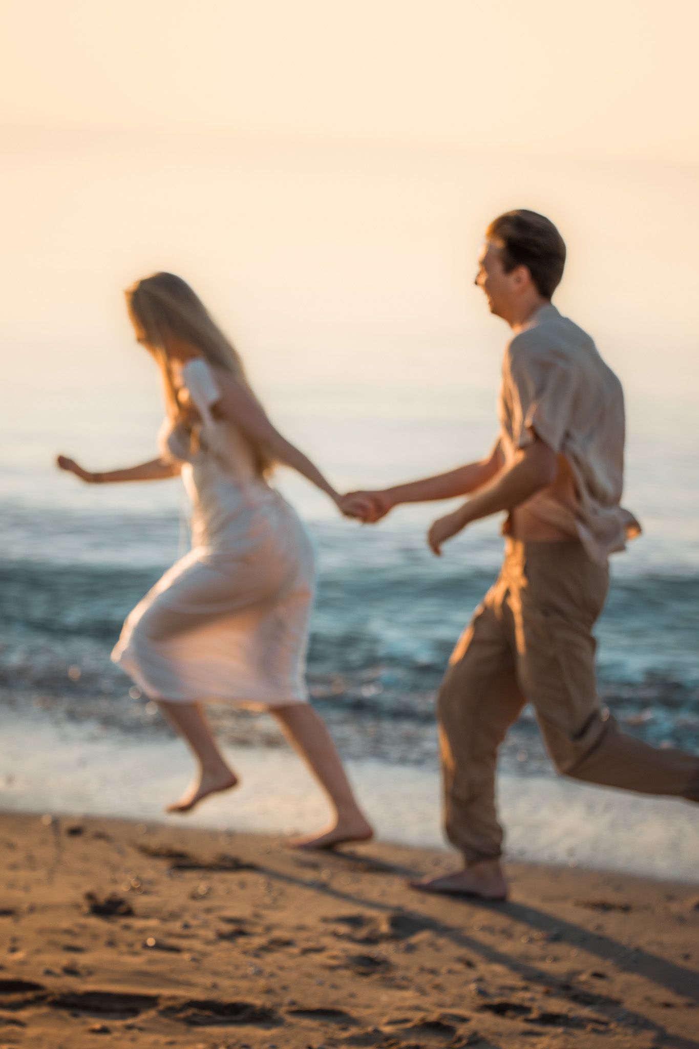 A couple playfully runs and dances together on a sunlit beach at sunset casting long shadows in the sand.