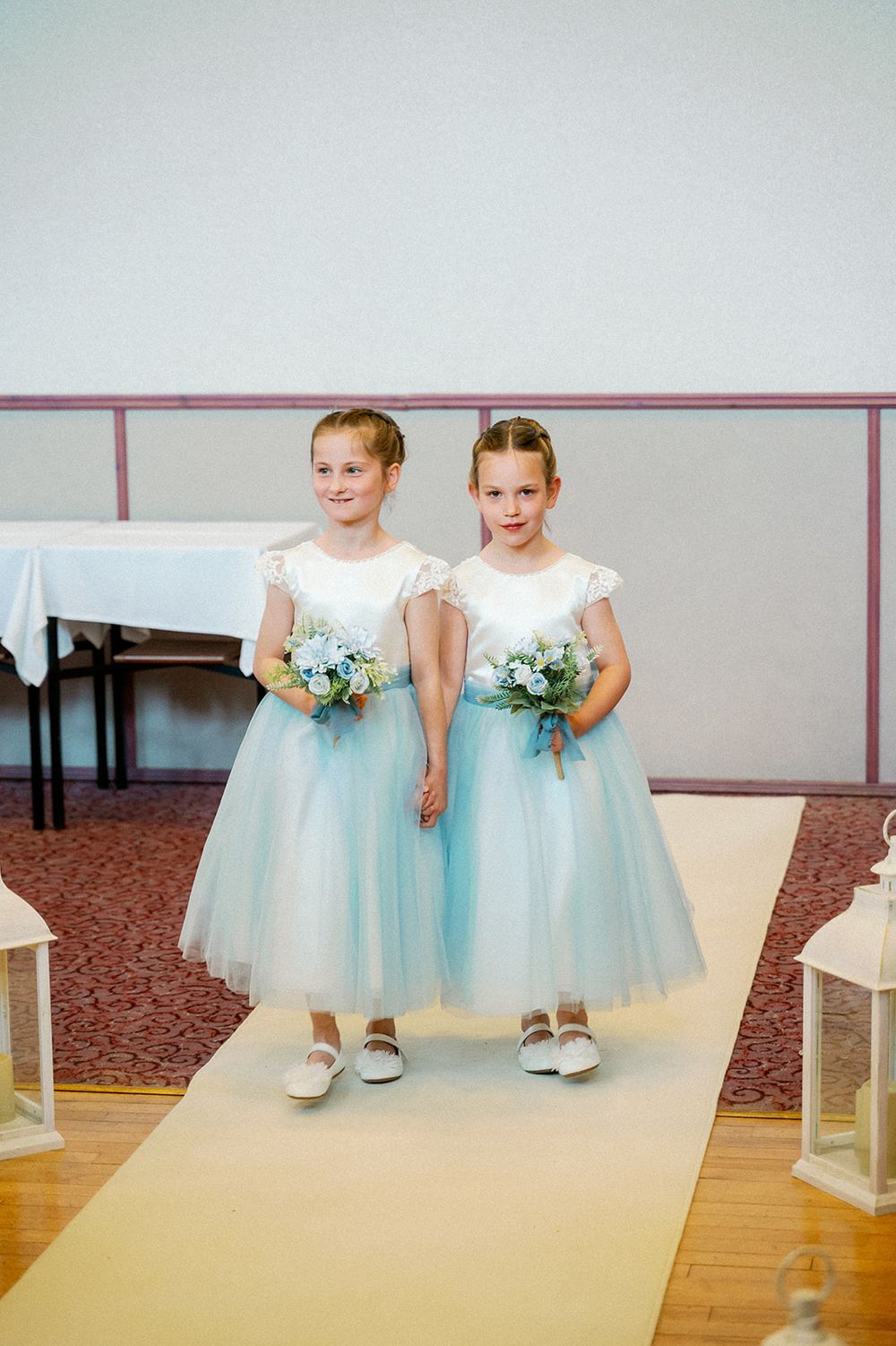 Two flower girls in matching blue and white tulle dresses hold bouquets on a gold aisle runner.