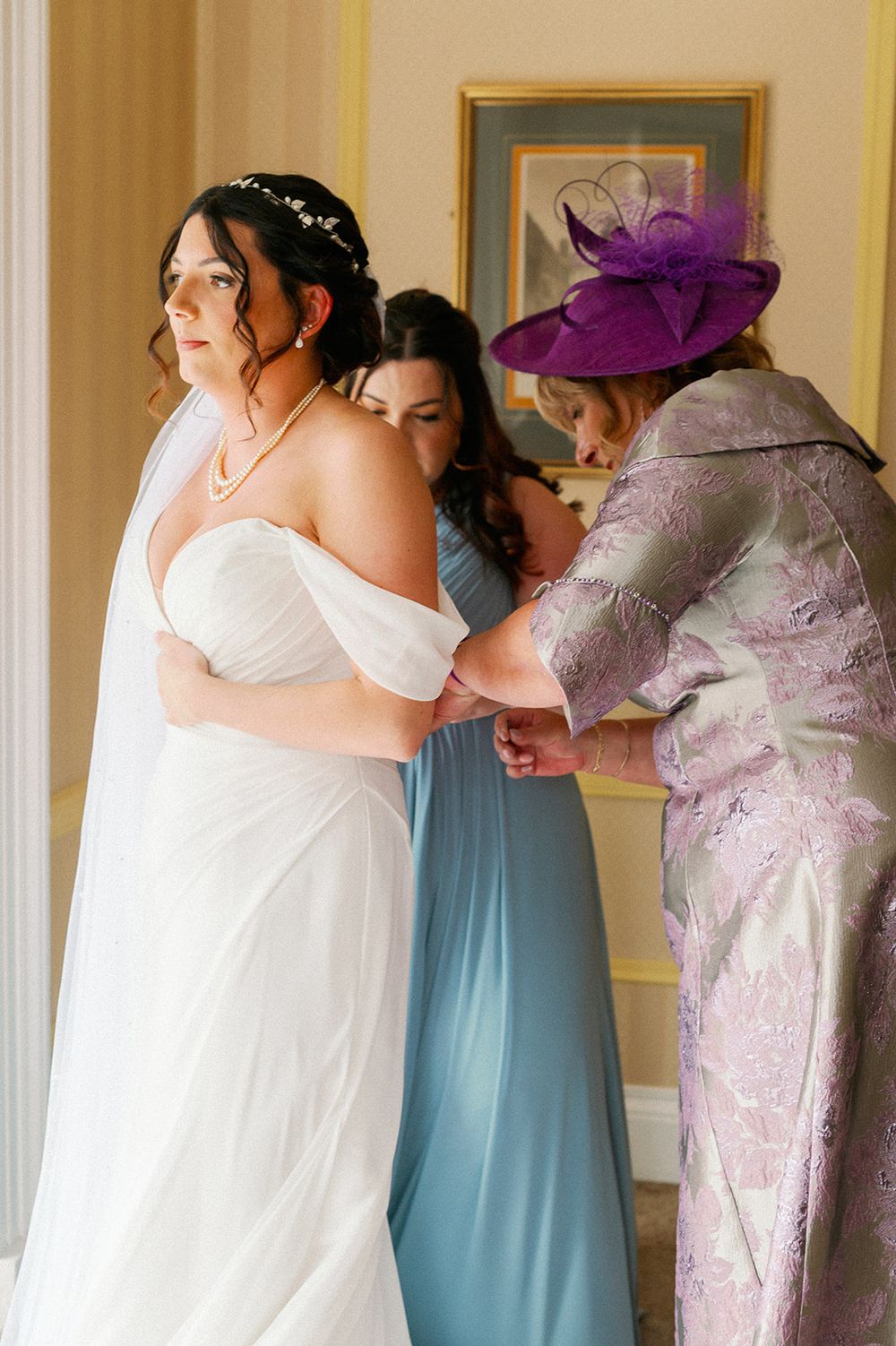 A wedding dress fitting scene with an elegant white gown and someone wearing a purple fascinator helping with adjustments.