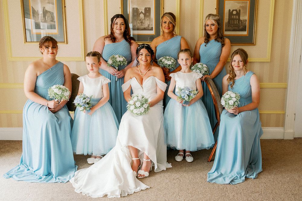 Bridal party in matching light blue dresses holding white bouquets pose together for a formal photo.