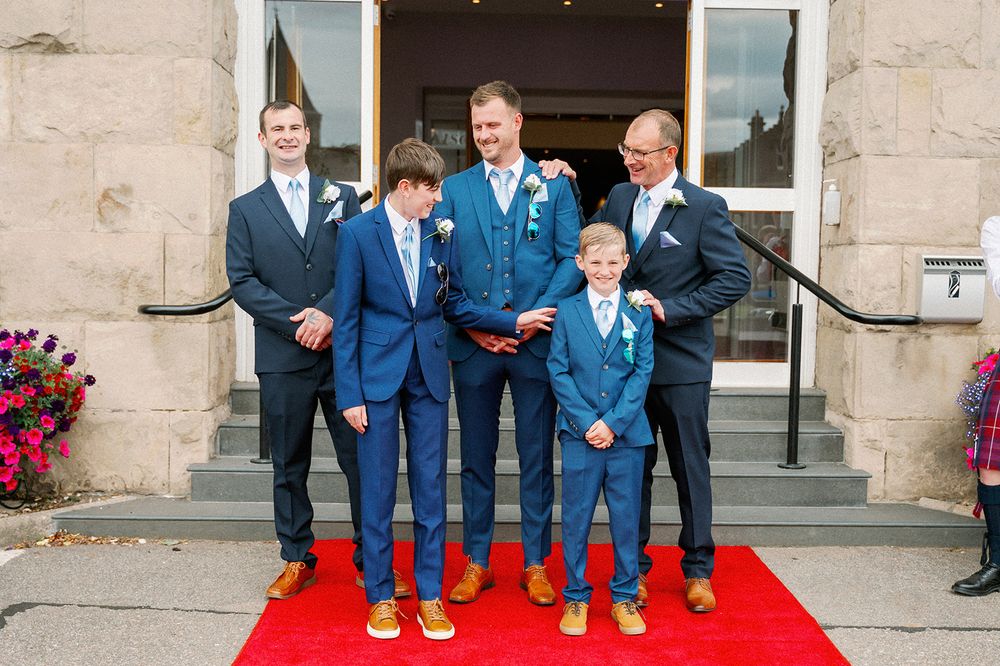 A group of grooms in matching blue suits standing together on stone steps and a red carpet at a Laichmoray wedding.