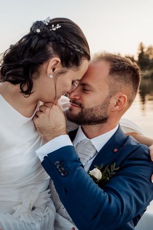Bride and groom sharing a romantic moment, touching foreheads, dressed in wedding attire near a serene lake at sunset.