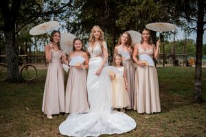 Bride in white gown poses with bridesmaids in champagne dresses holding parasols outdoors under trees.
