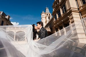 Couple sharing a romantic kiss outdoors with a flowing wedding veil, elegant architecture and blue sky in the background.