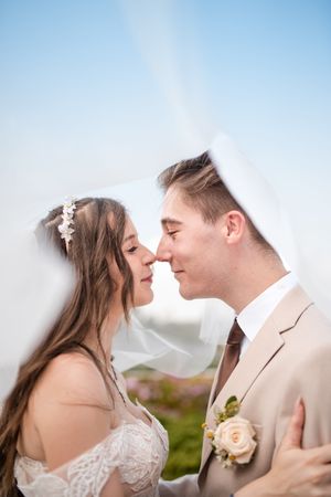 Bride and groom sharing a romantic moment under a wedding veil against a blue sky backdrop.