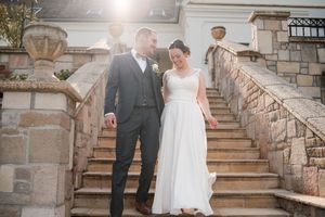 Bride and groom walking down stone steps at elegant venue, bride in white gown, groom in gray suit.