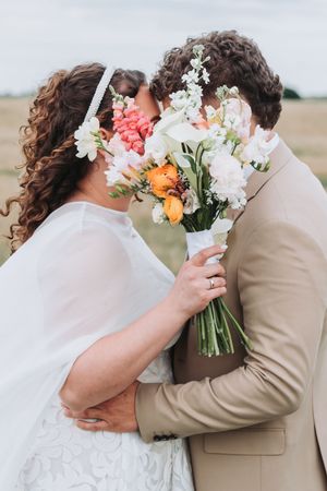 Bride and groom kissing behind a colorful wedding bouquet of white, orange, and pink flowers in an outdoor field.