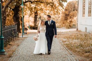 Bride and groom walk hand-in-hand on cobblestone path surrounded by autumn trees in golden light.