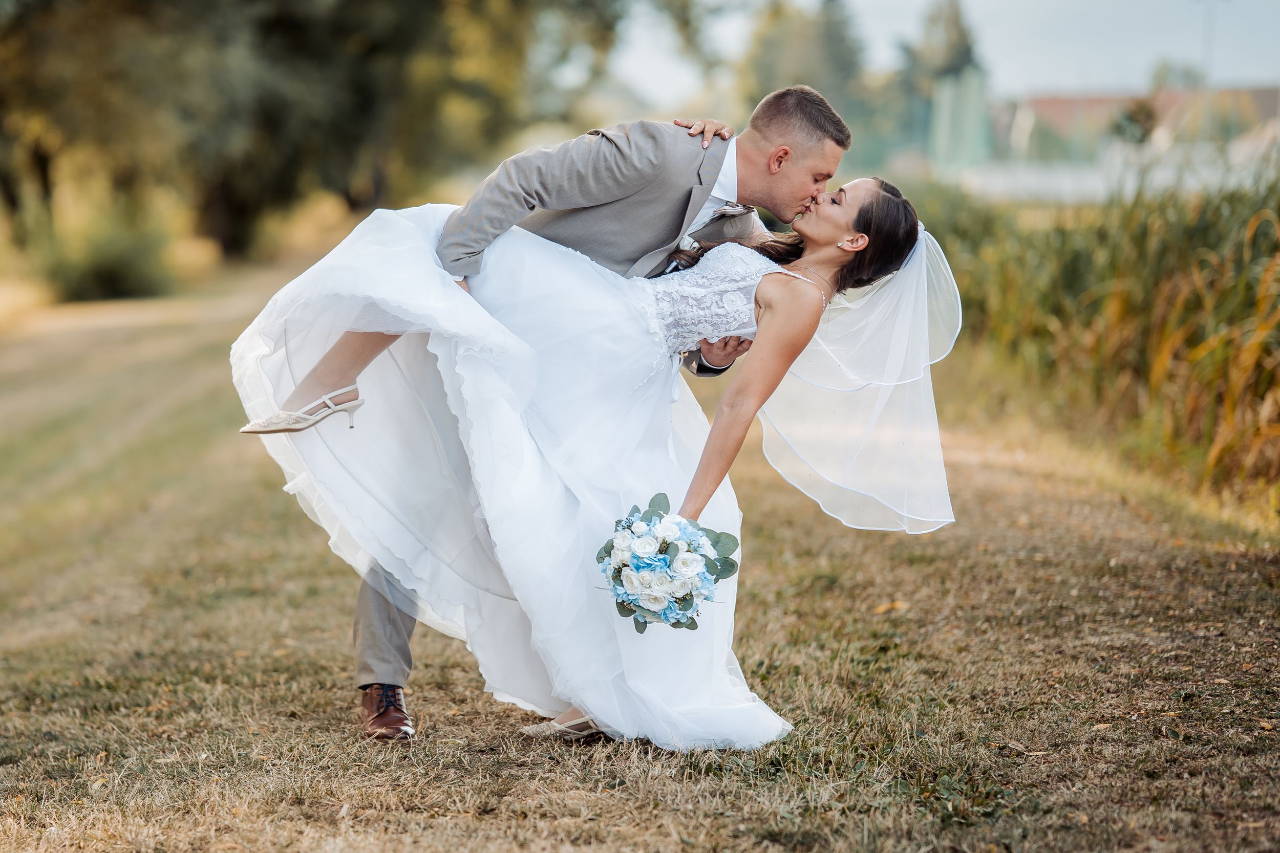 Groom in gray suit dips bride in white wedding dress for a romantic kiss outdoors in golden sunlight.