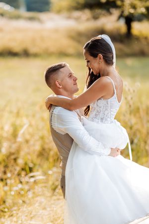 Groom lifts smiling bride in white lace wedding dress in a sunny golden field, gazing lovingly.