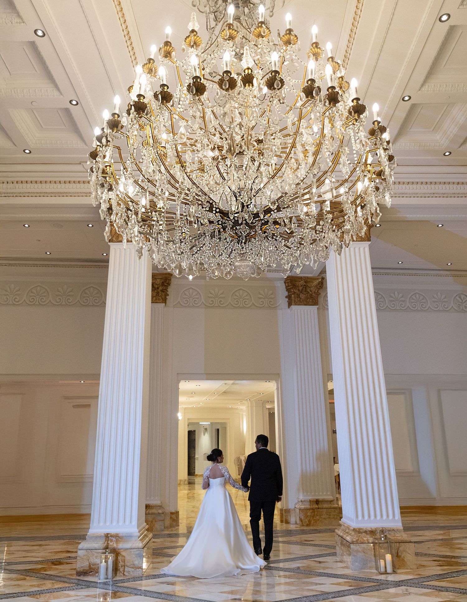 Wedding couple walks hand-in-hand beneath a grand crystal chandelier in an elegant ballroom.