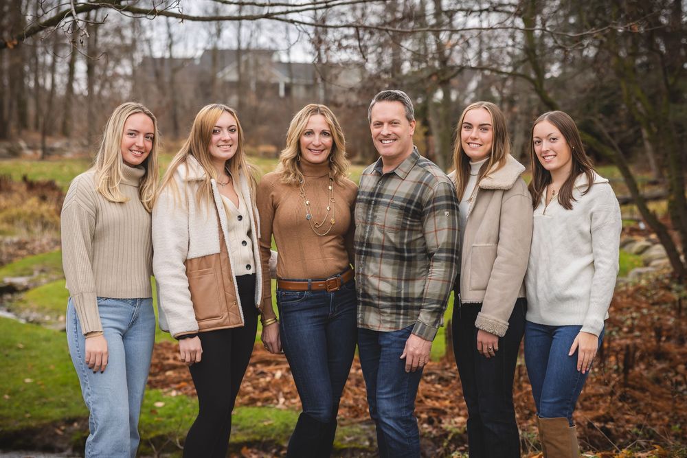 A group poses together outdoors in casual fall attire with sweaters and jeans against an autumn forest backdrop.
