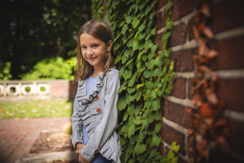 A young person stands by a brick wall with climbing ivy, wearing a gray long-sleeved shirt and smiling.