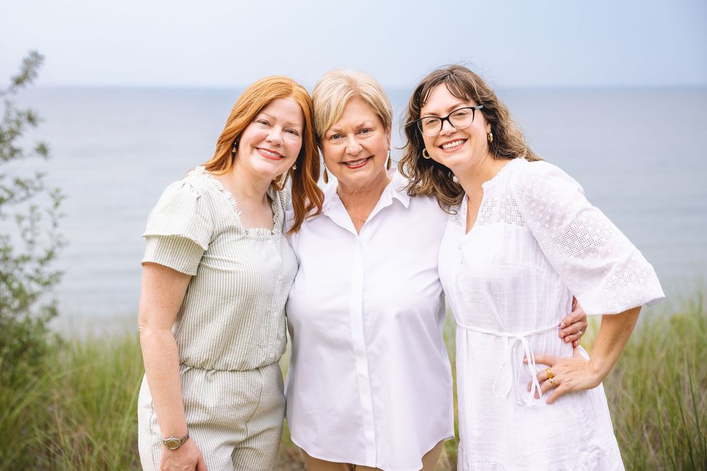 A family group poses together outdoors in coordinating white and pink outfits against a coastal landscape background.