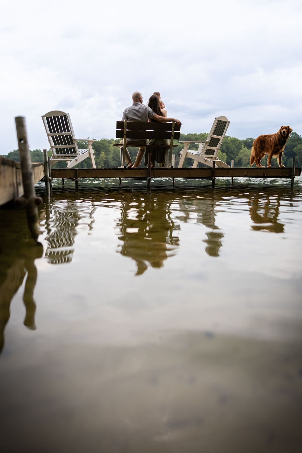 Silhouettes of people and chairs reflected in calm water at a dock during sunset.