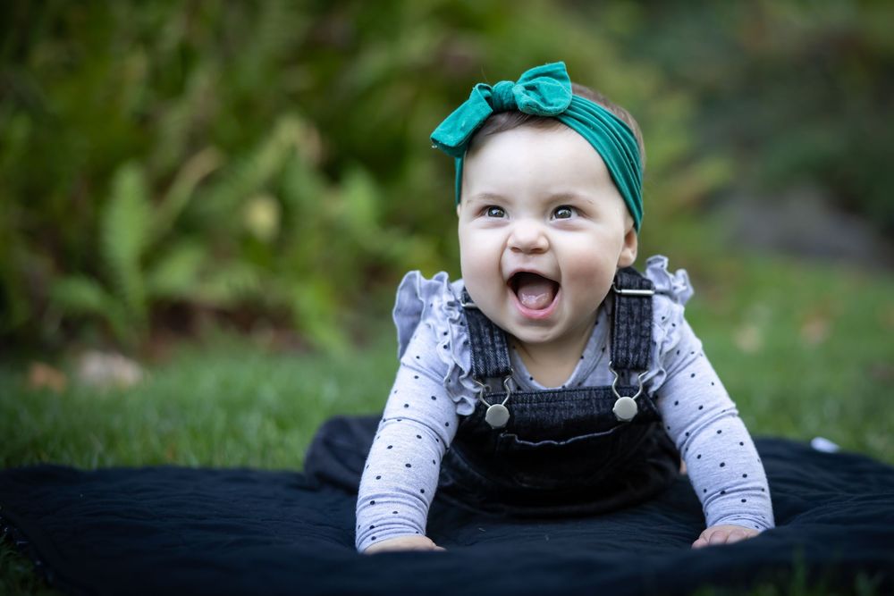 Happy baby wearing teal bow headband and white outfit smiling at camera.