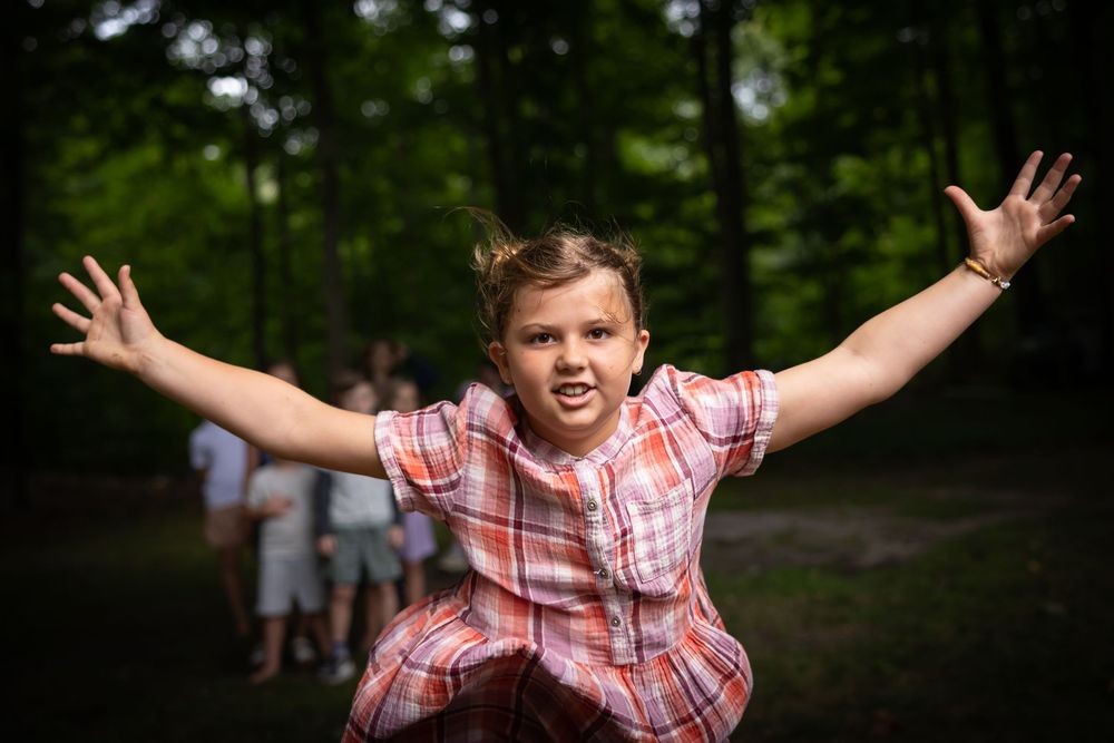 A person in a red and white plaid shirt jumps joyfully in a wooded outdoor setting.