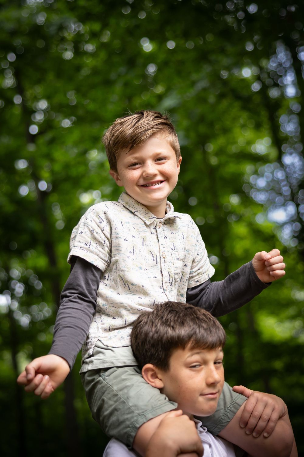 Two children playing outdoors with one sitting on the other's shoulders against a blurred green background.