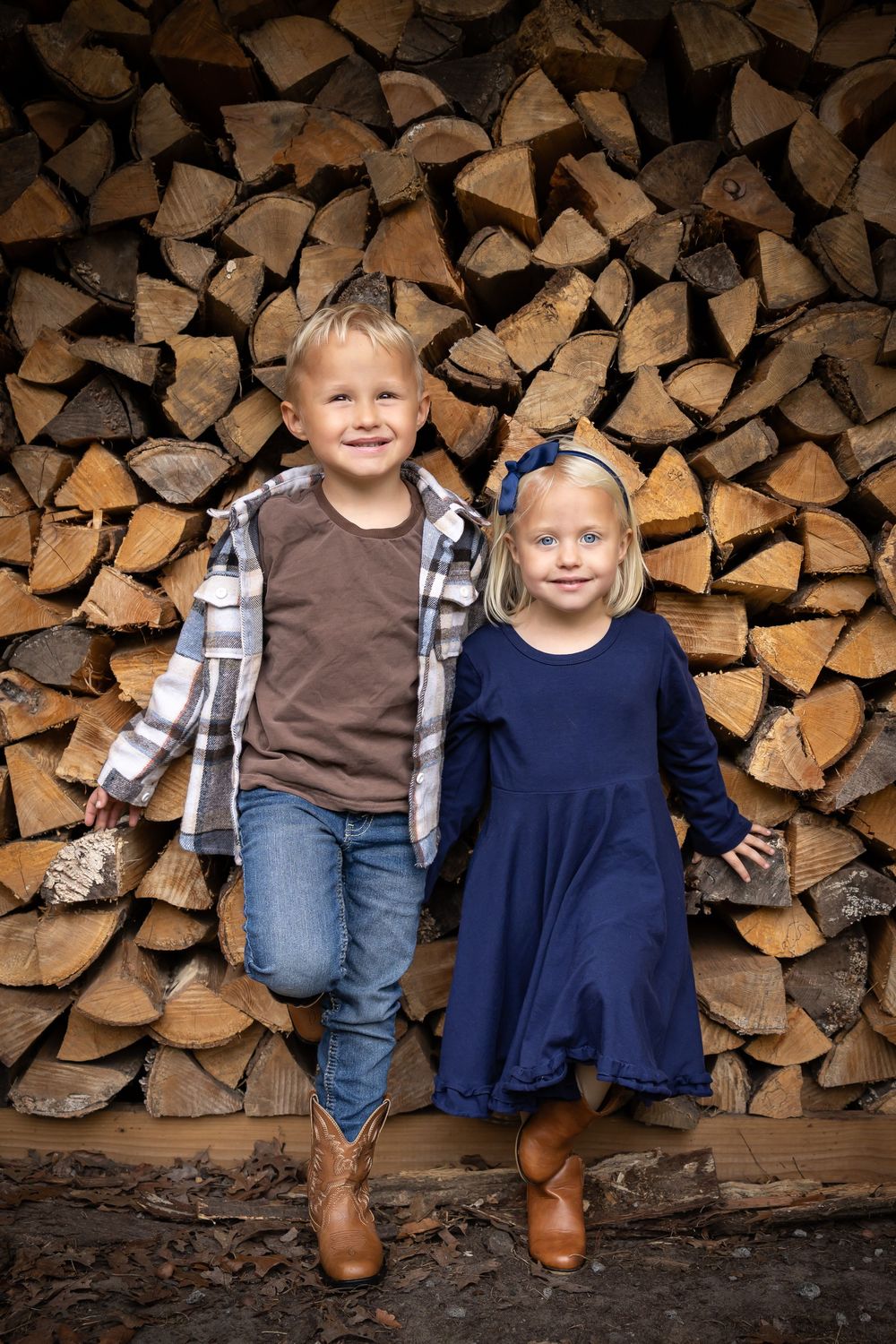 Two children stand together in front of a rustic woodpile wearing coordinated casual outfits.