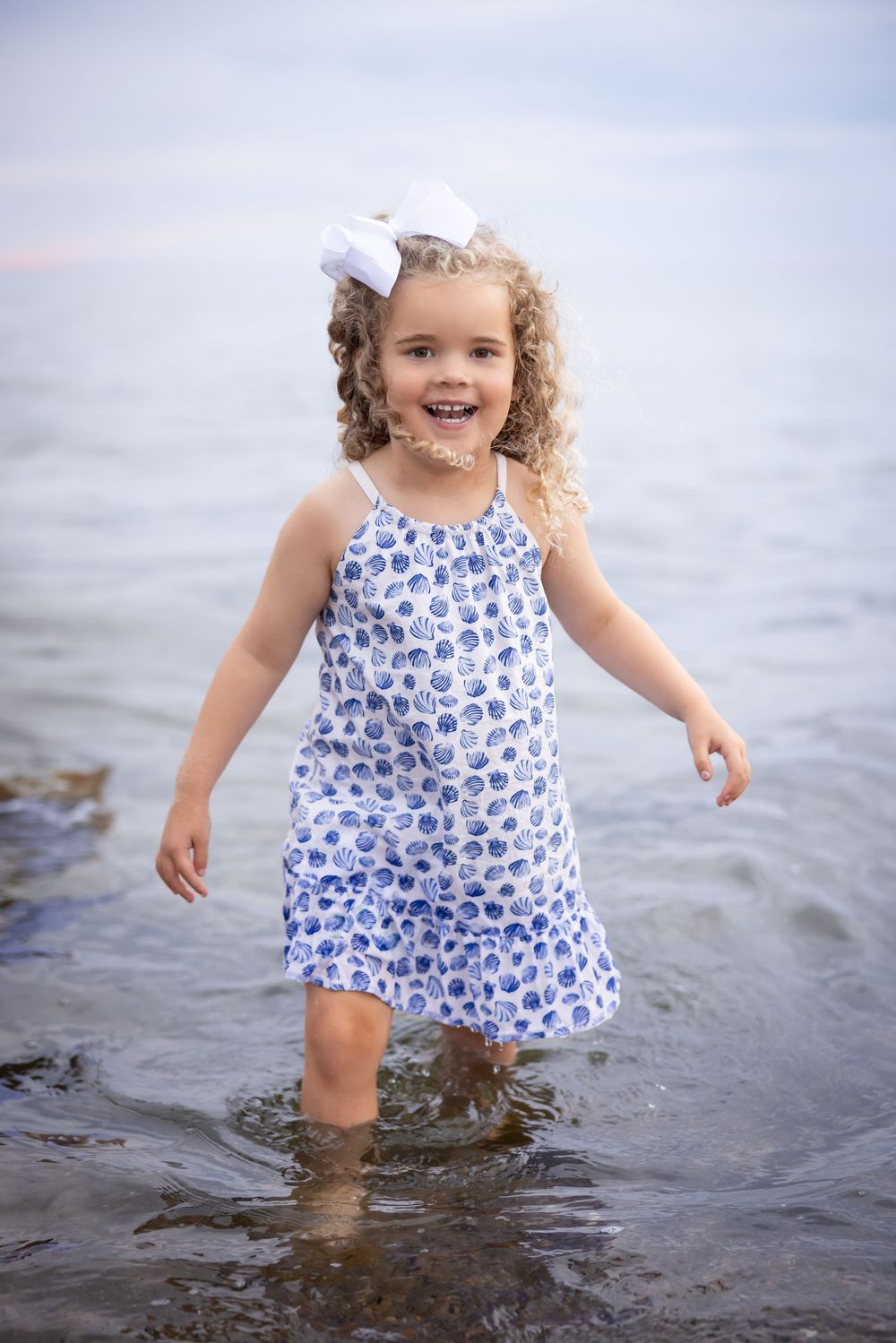 A child wearing a blue and white floral dress splashes joyfully in shallow ocean water.