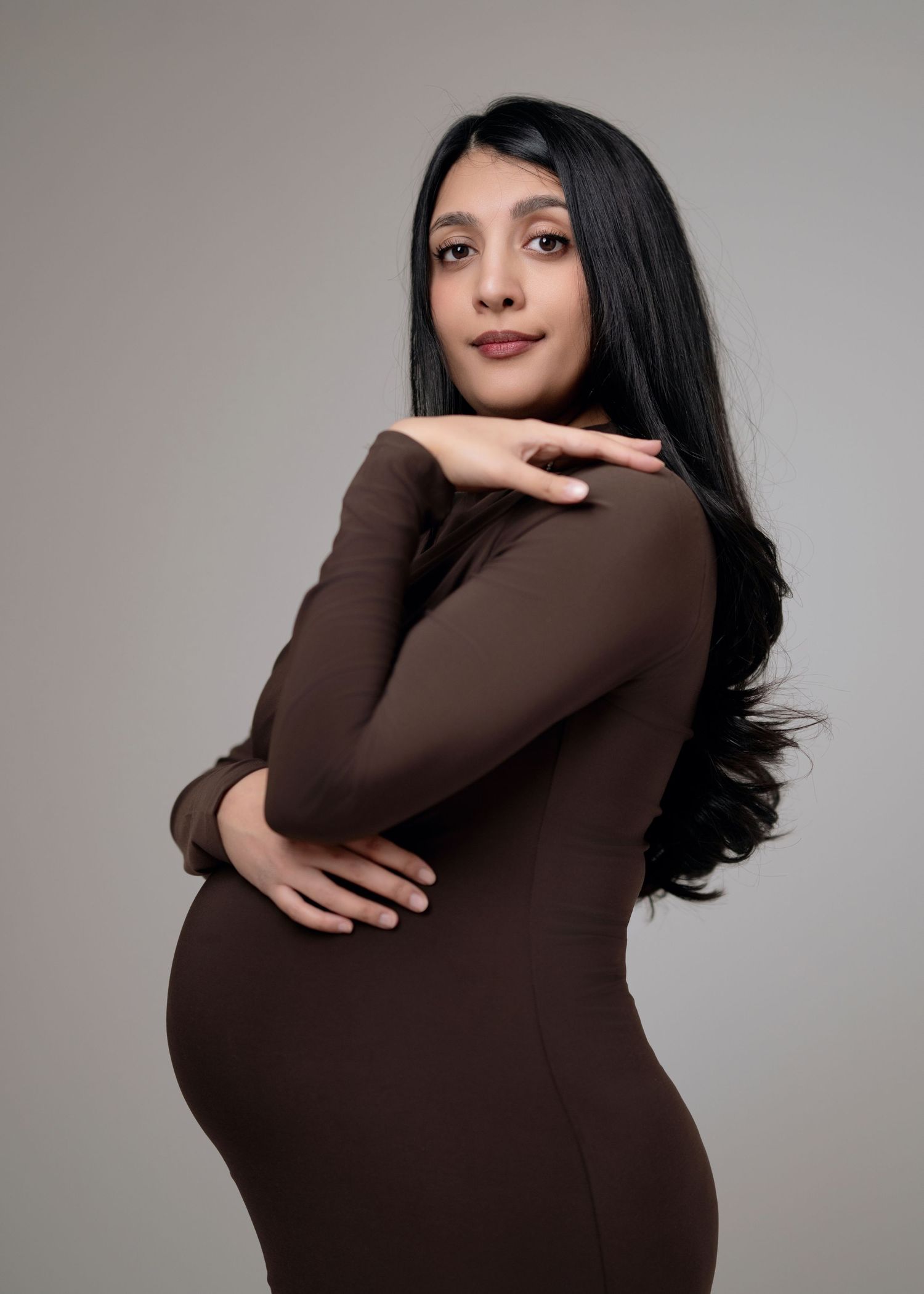 A pregnant person in a fitted brown dress poses against a gray background in a maternity photoshoot.