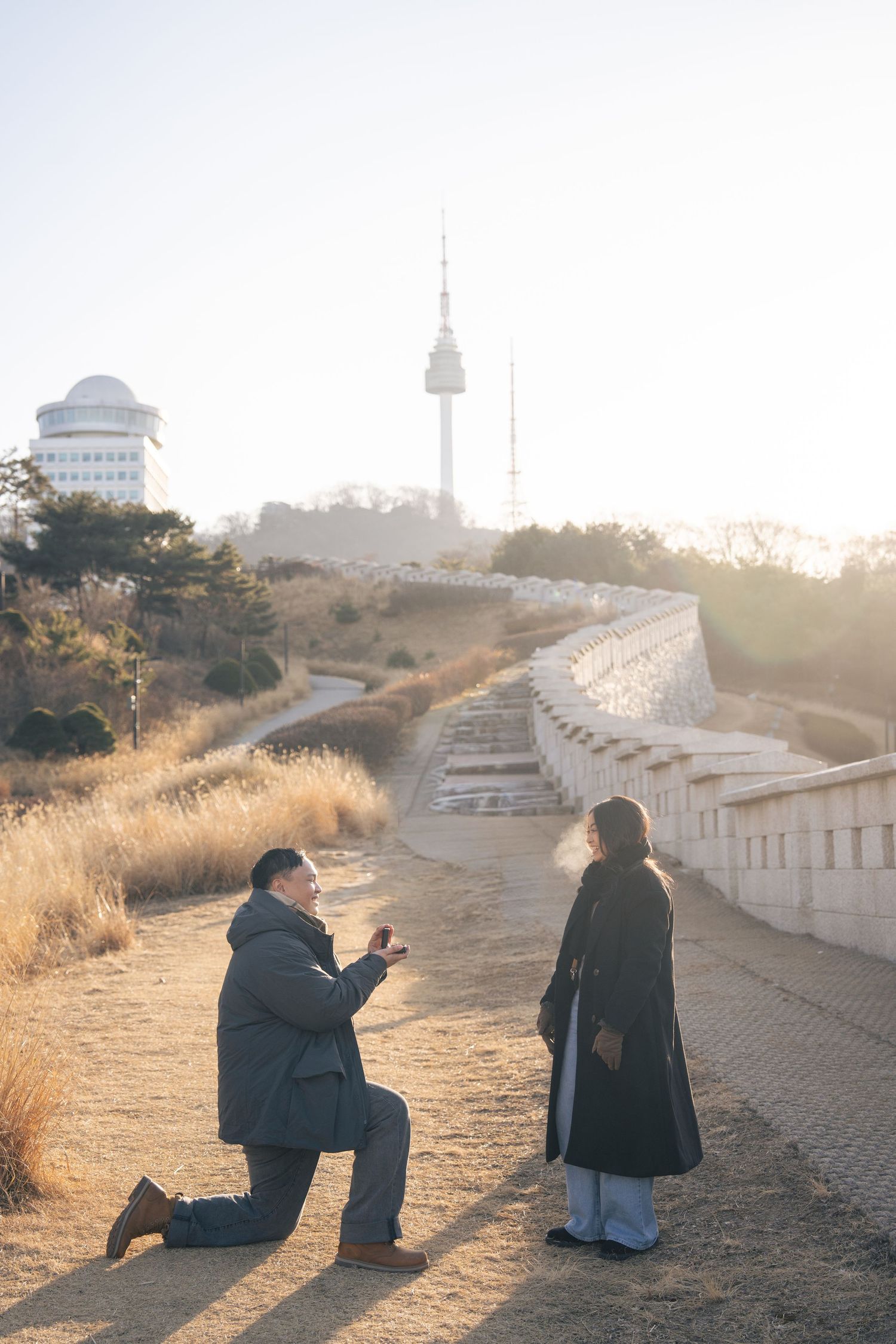 Two people sit together on a stone path at sunset with a lighthouse and buildings visible in the misty background.