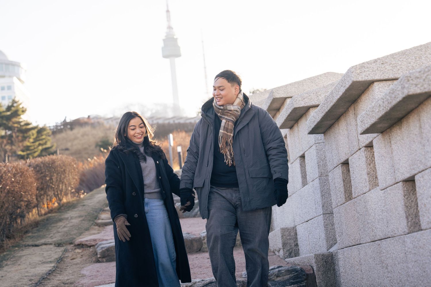 A couple walks together along a stone wall path with N Seoul Tower visible in the background on a winter day.