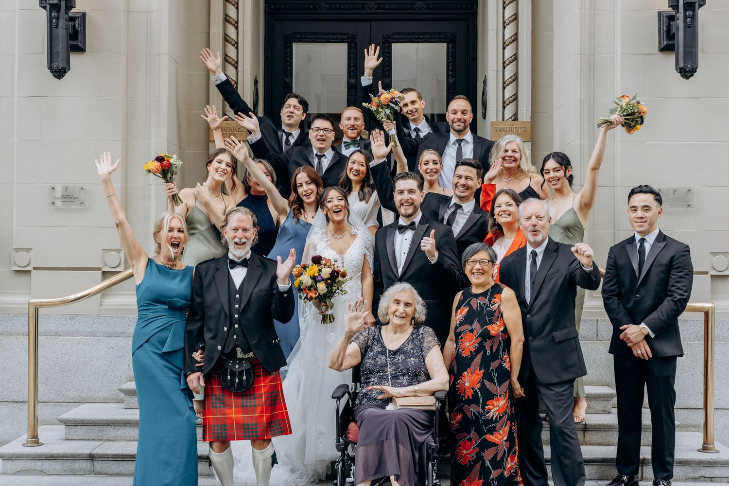 A large group of wedding guests gathered on stone steps celebrate joyfully with raised arms and smiles.