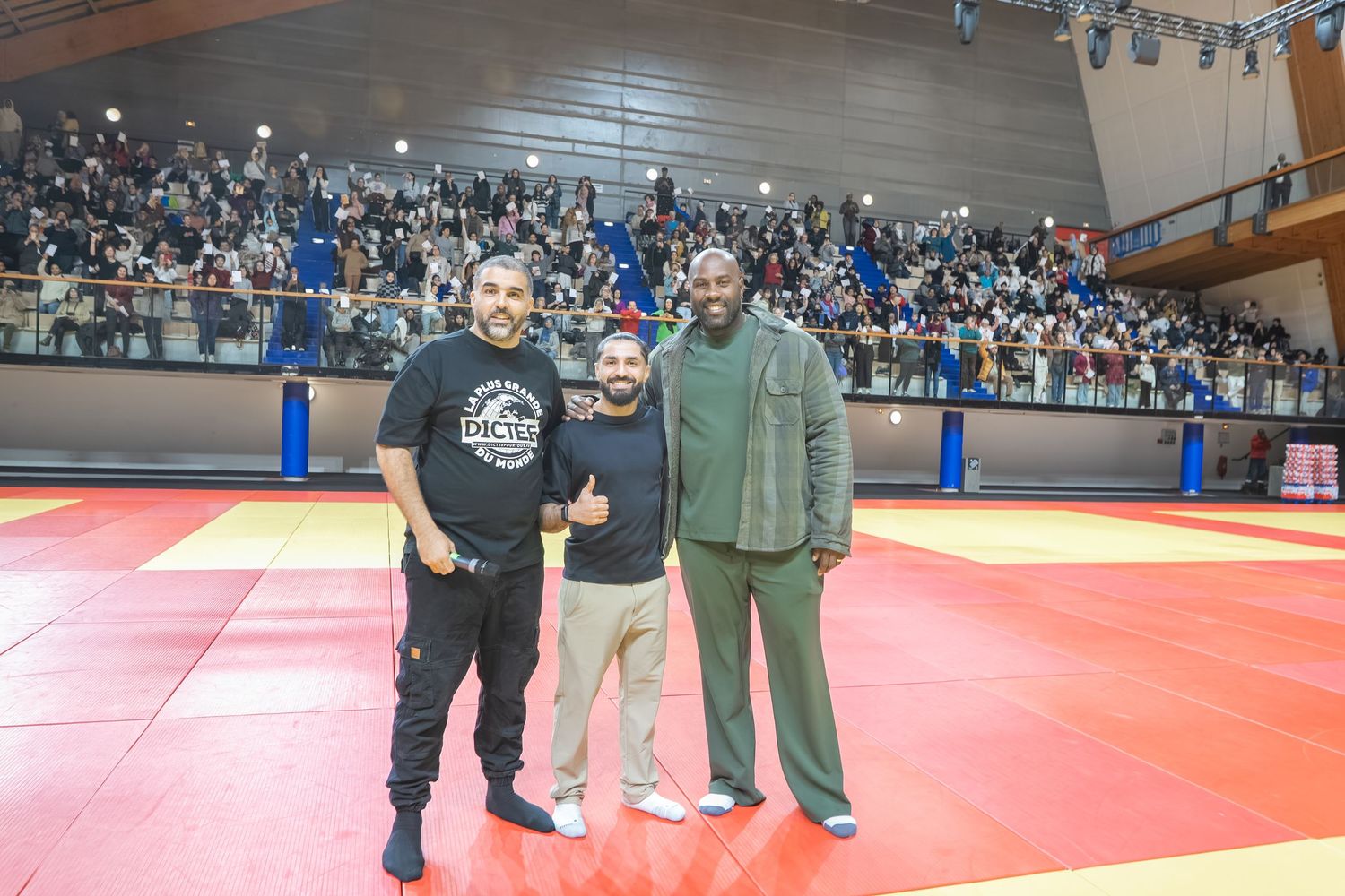A group of people stand on red wrestling mats in a gymnasium during what appears to be a sporting event.