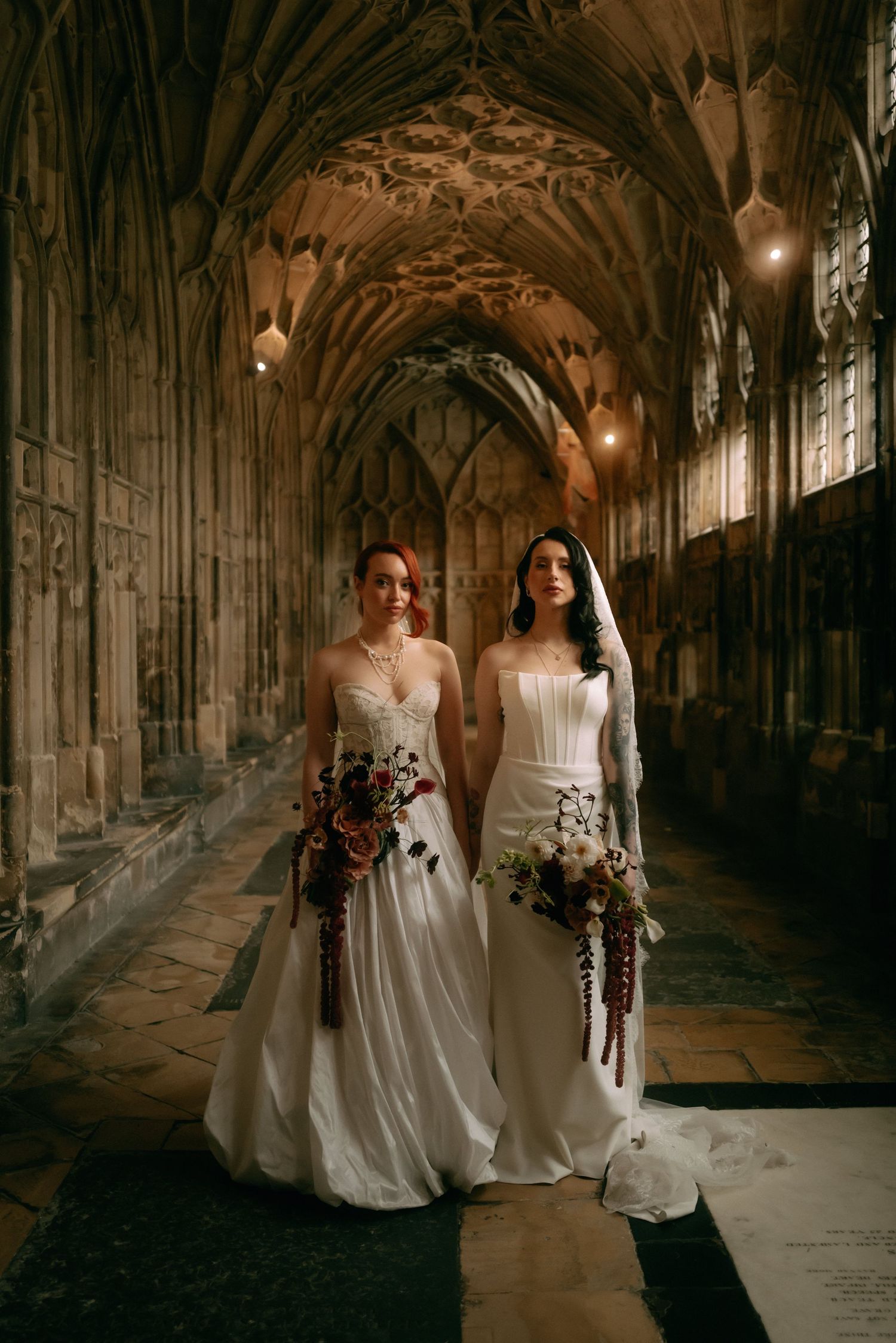 Two brides in elegant white wedding dresses pose together in a dramatic gothic cathedral corridor with stone archways.