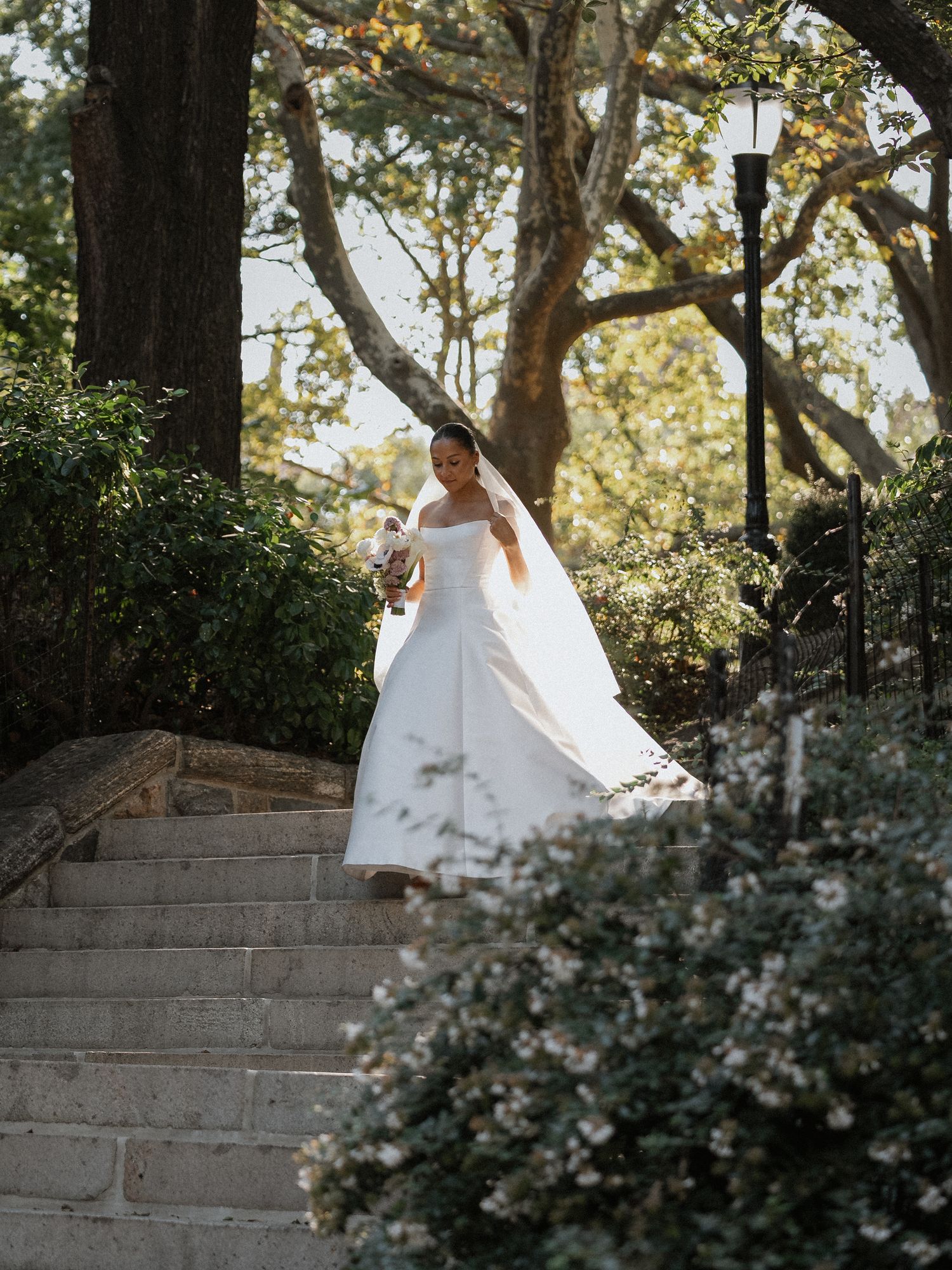 A bride in a flowing white wedding dress descends stone garden steps surrounded by lush greenery and dappled sunlight.