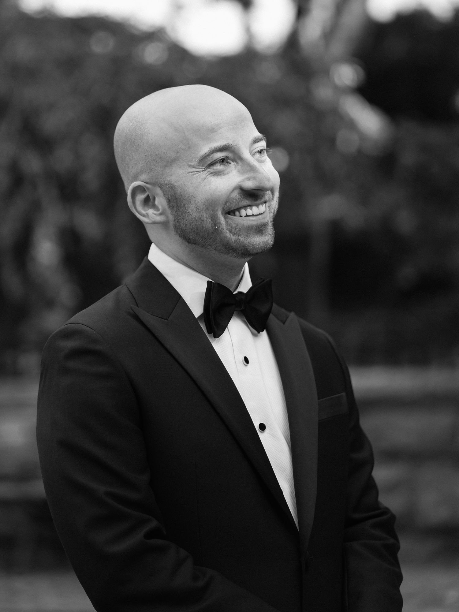 Black and white portrait shows groom in tuxedo smiling during outdoor wedding.