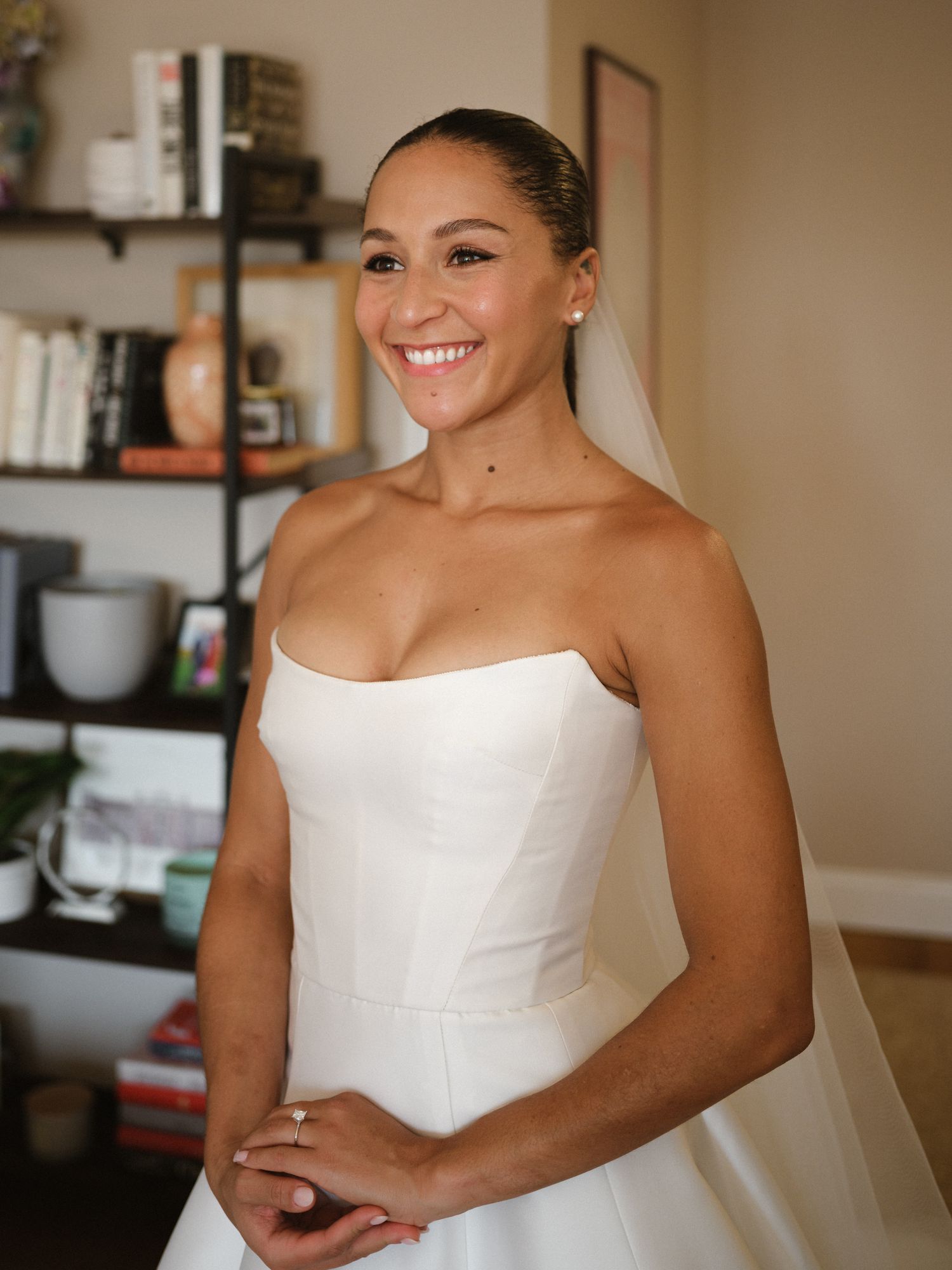 A bride wearing a classic white strapless gown smiles while getting ready in a sunlit room.