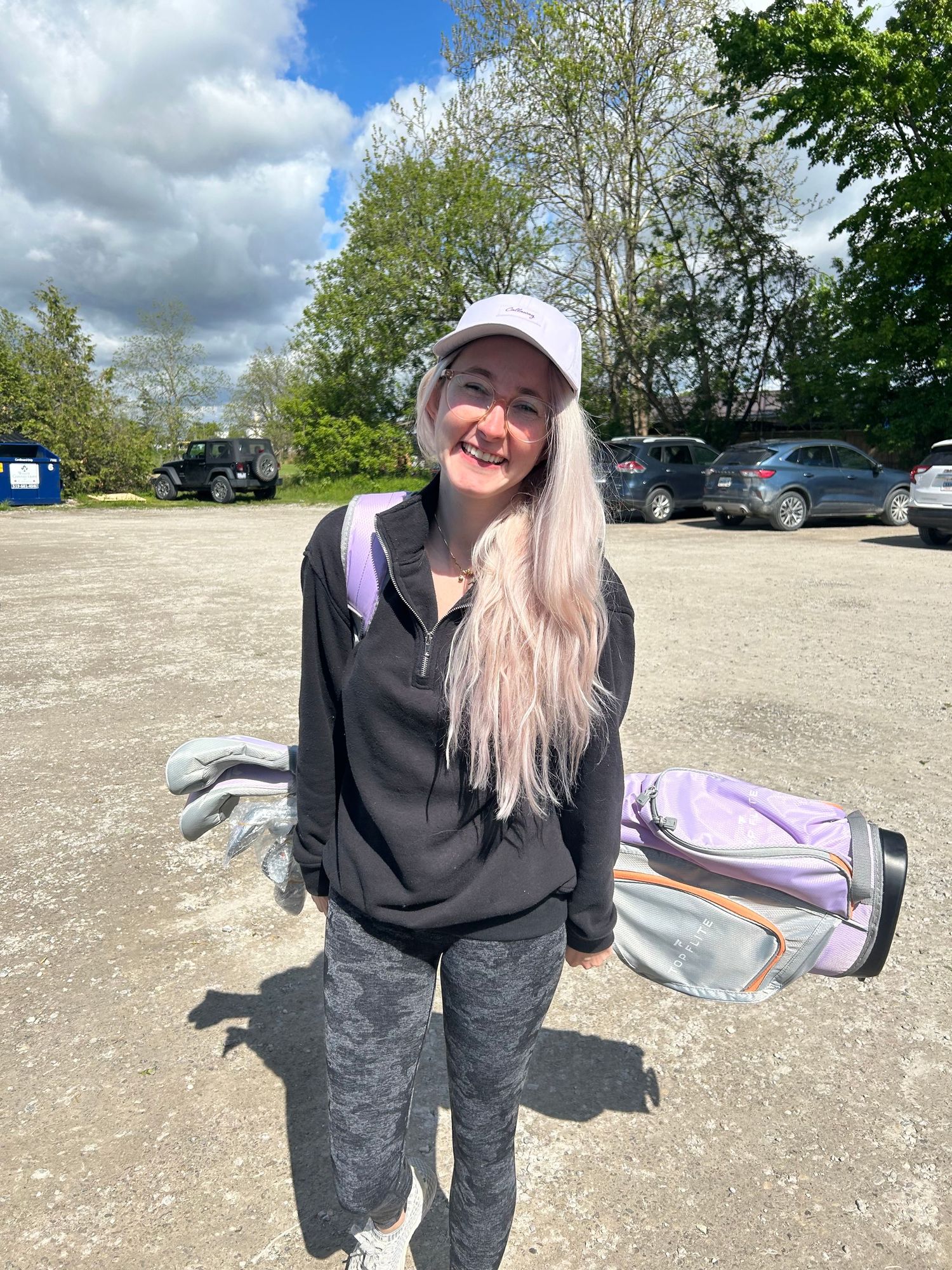 A person in black overalls and a baseball cap poses outdoors in a gravel parking area on a sunny day.