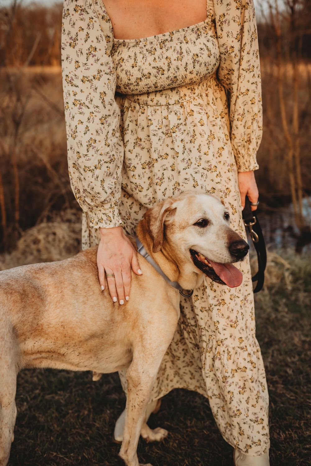Woman in floral dress standing with a yellow Labrador Retriever on a leash outdoors at golden hour.