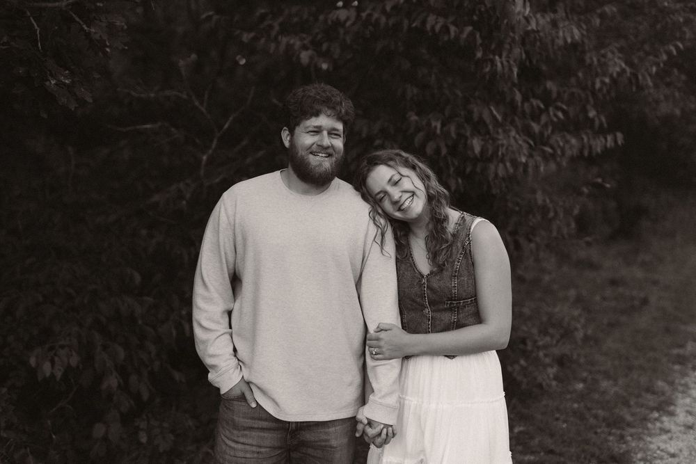 A romantic couple embraces while standing against a dark wooded background.