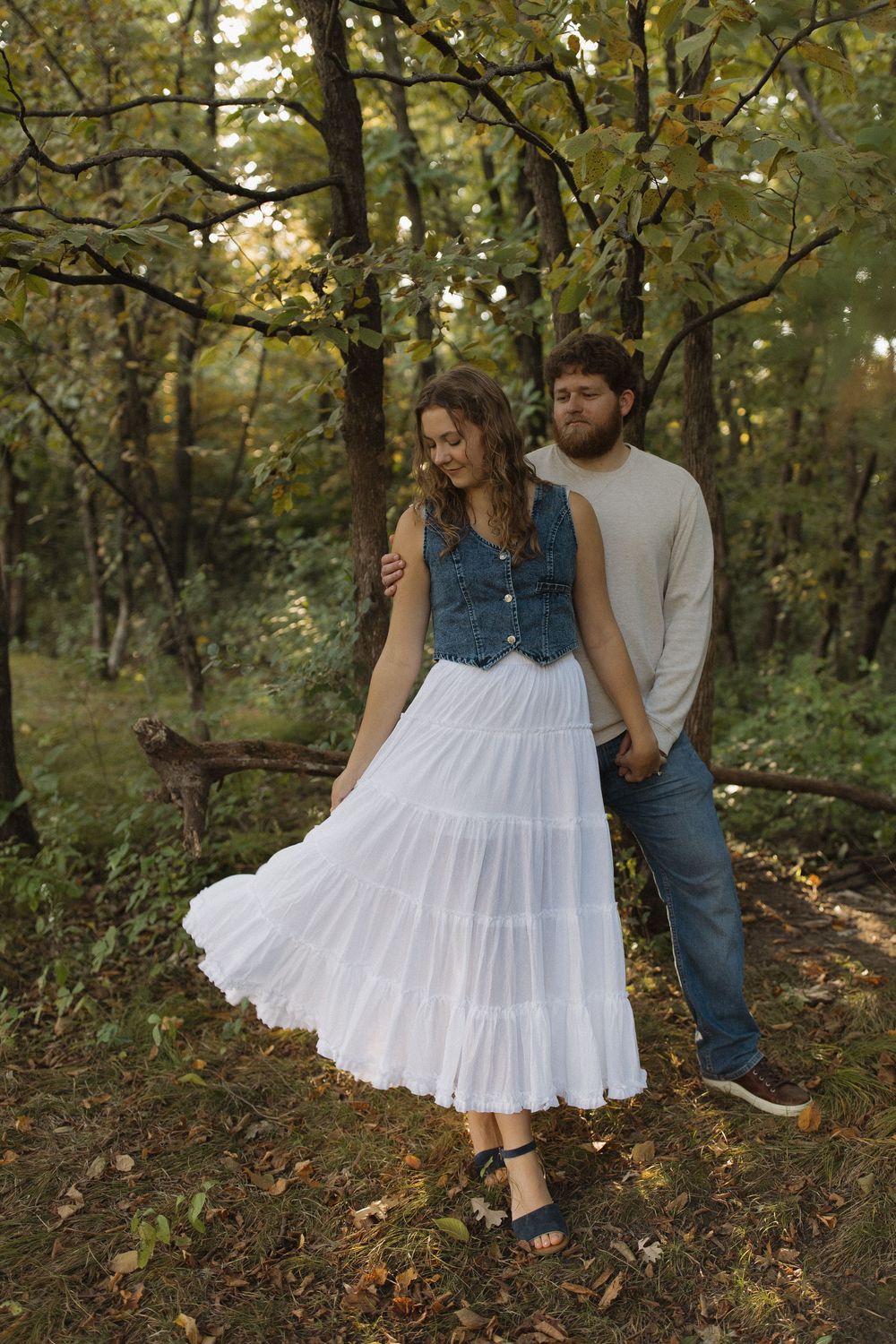 A couple sits together on a hillside wearing casual clothes among autumn trees.