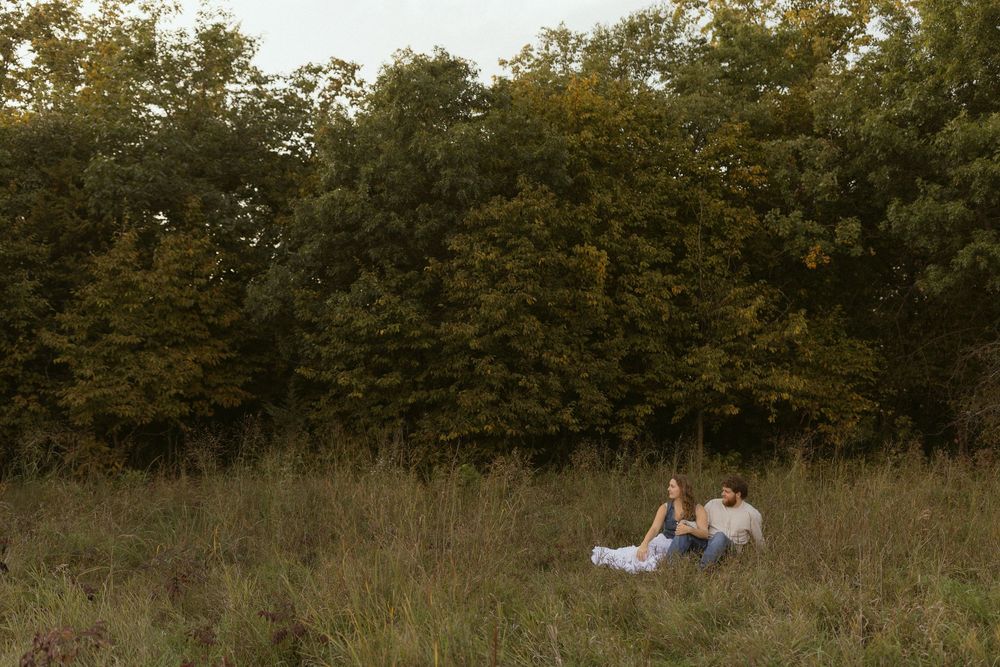 A distant view of a couple sitting in a grassy field surrounded by trees.