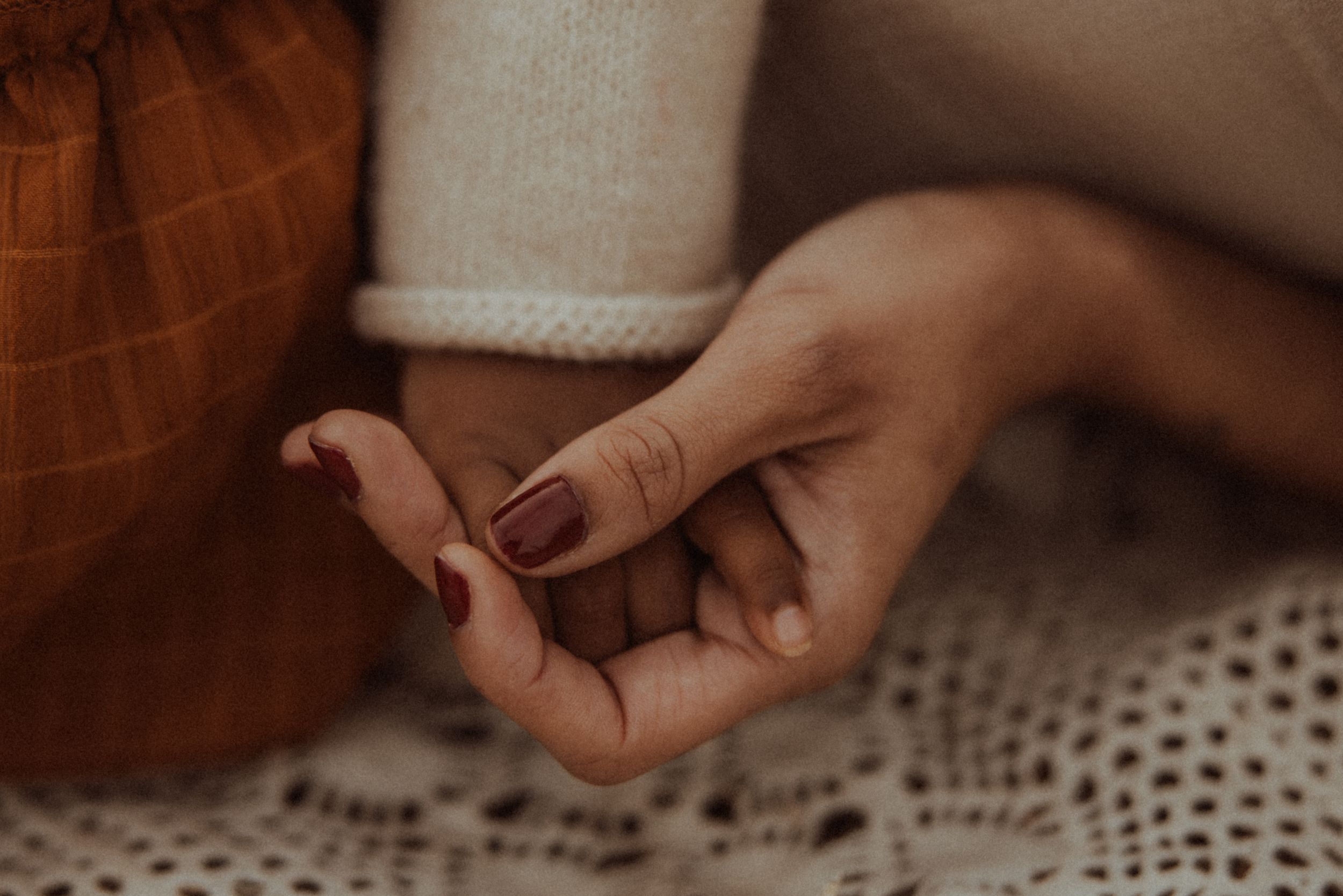 Close up of hands with burgundy nail polish resting on white lace fabric in warm lighting.