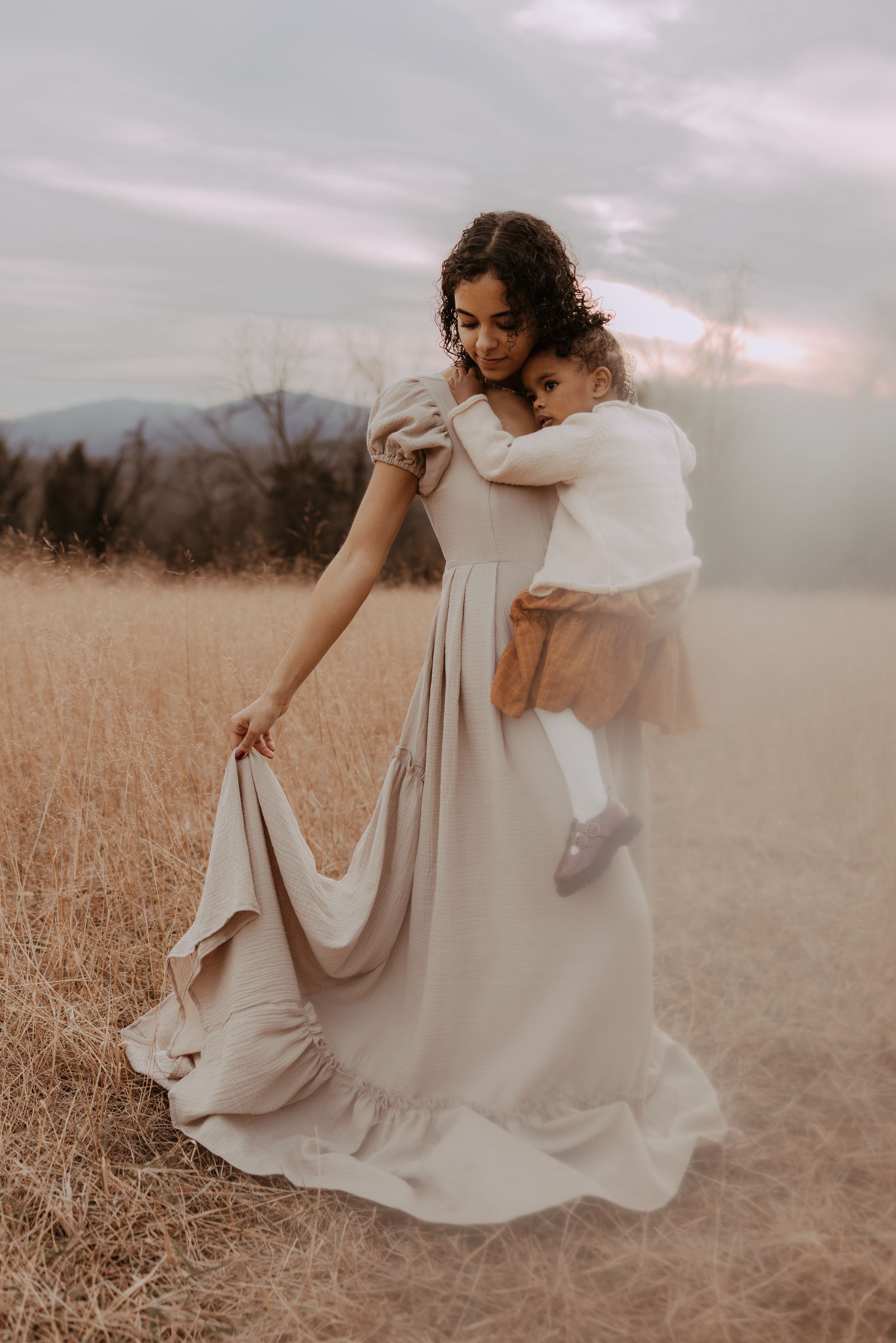 Two figures in flowing white dresses embrace while dancing in a golden wheat field at sunset.