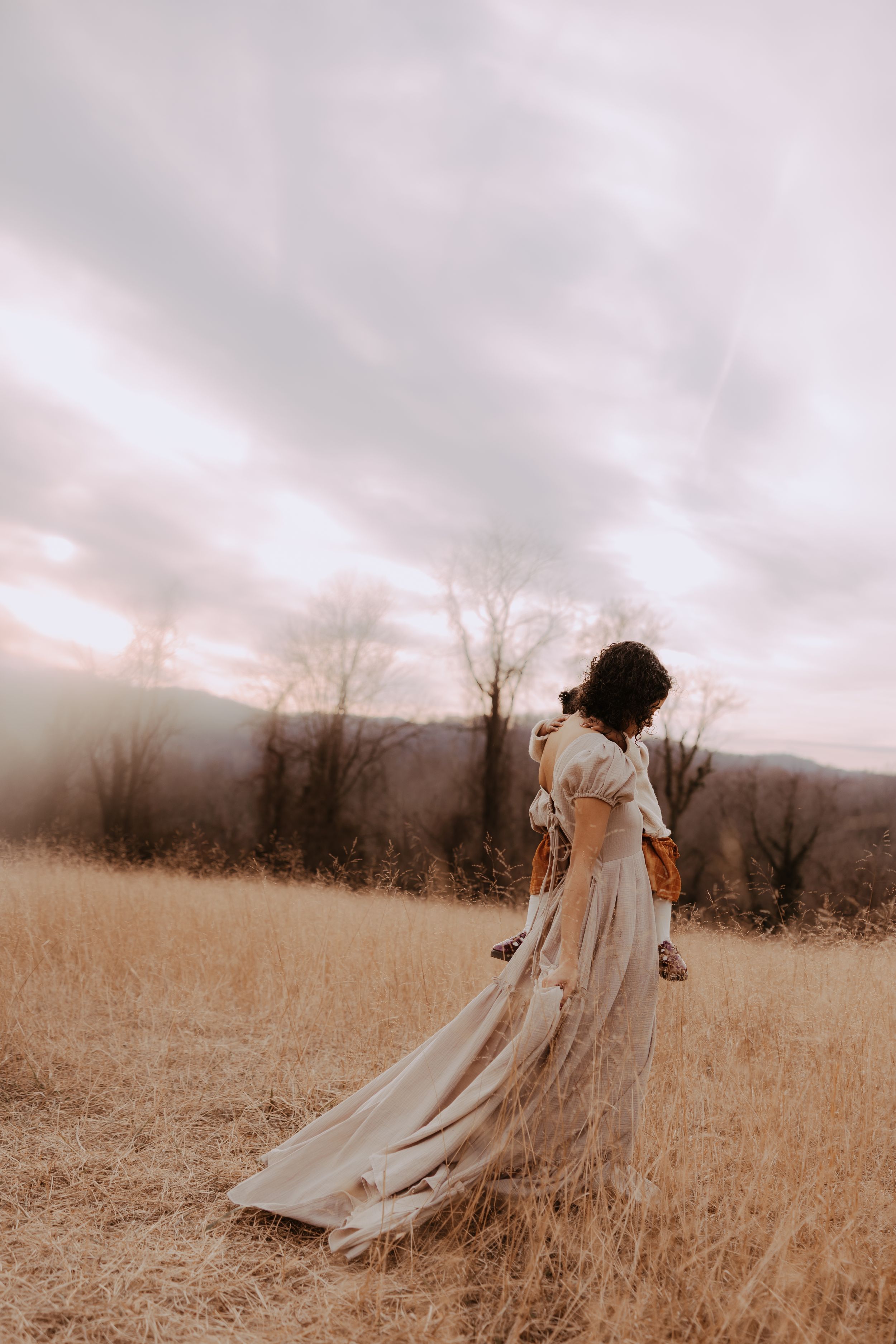 A dreamy figure in a flowing vintage dress walks through a sepia-toned autumn field under moody skies.