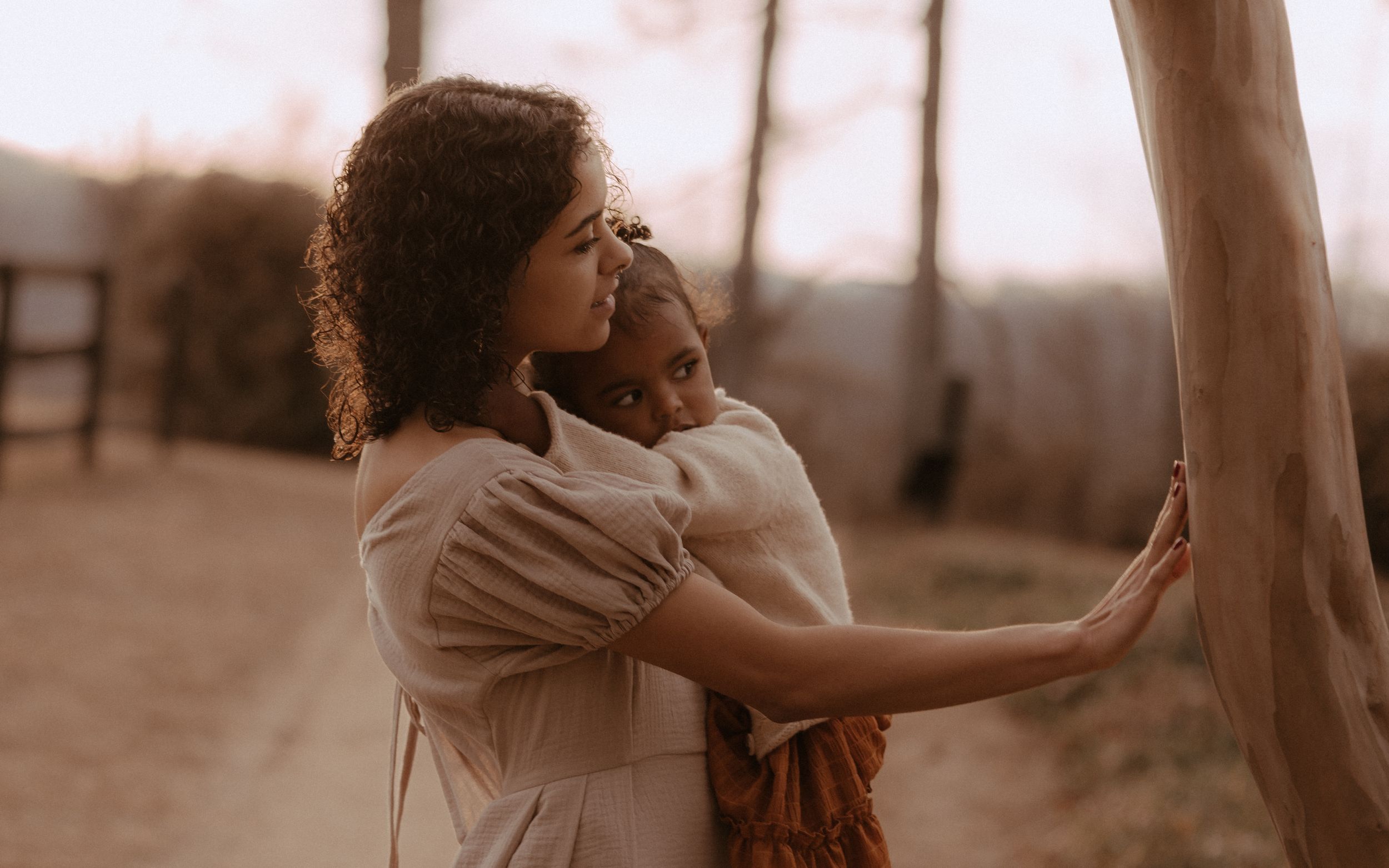 Two people hold each other close while standing near a tree trunk in sepia lighting.