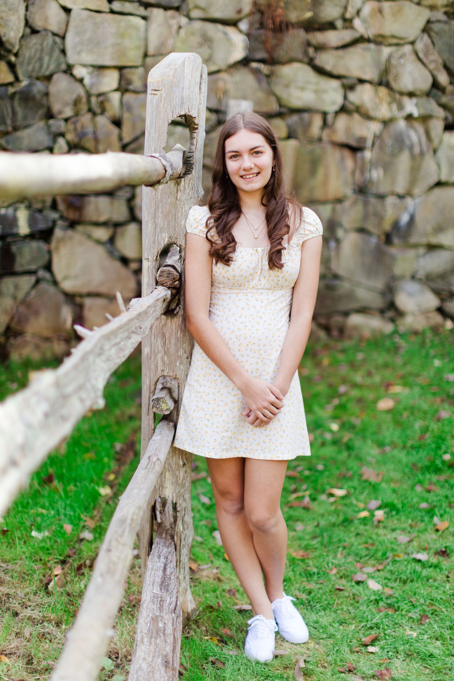 Senior in High school in floral dress smiling by rustic wooden fence with stone wall backdrop on green lawn for her Senior Photos