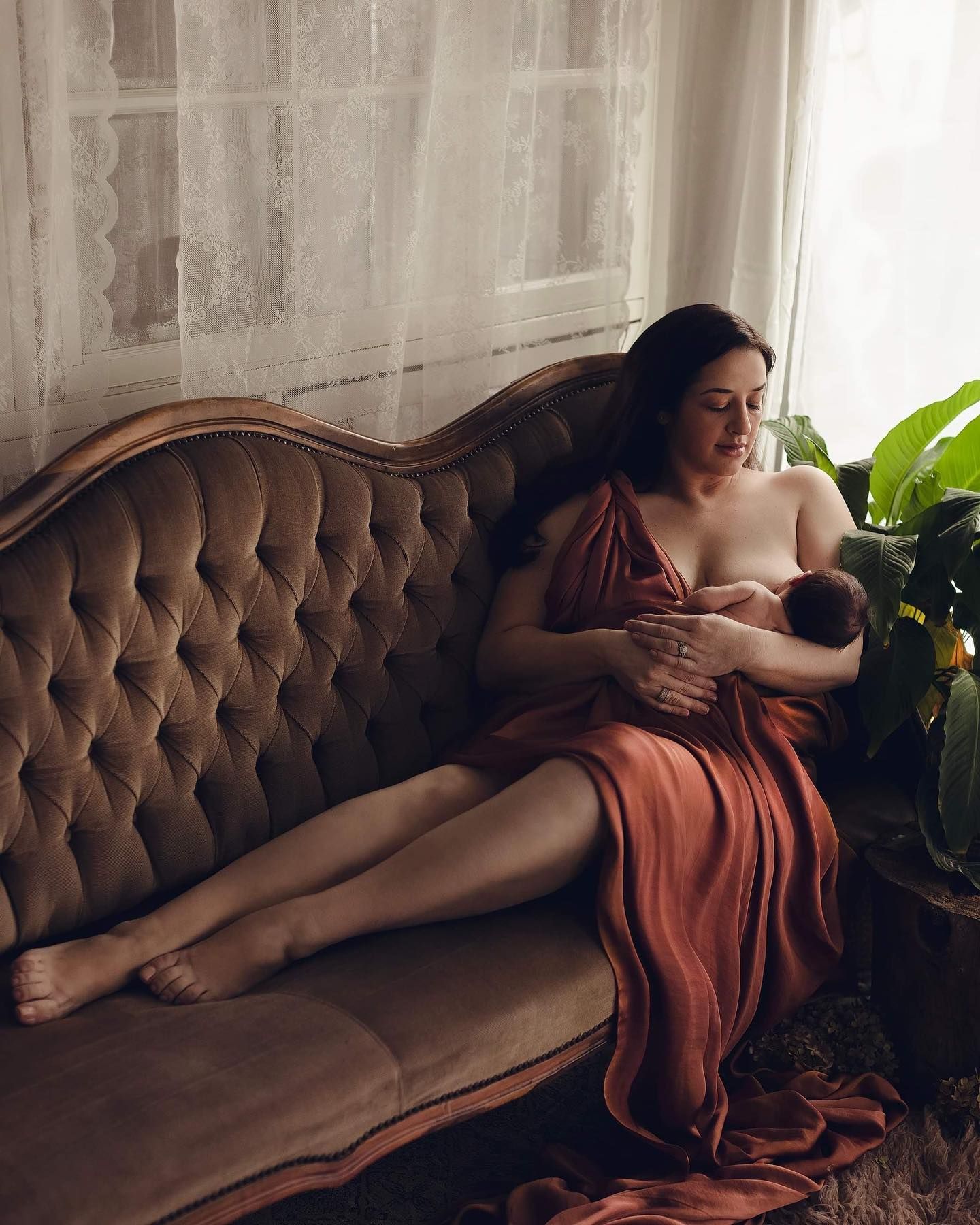A figure in a rust-colored dress reclines gracefully on a brown tufted sofa near indoor plants in soft natural light.