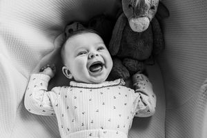 Black and white photo of a laughing baby lying next to a stuffed donkey toy on a blanket.